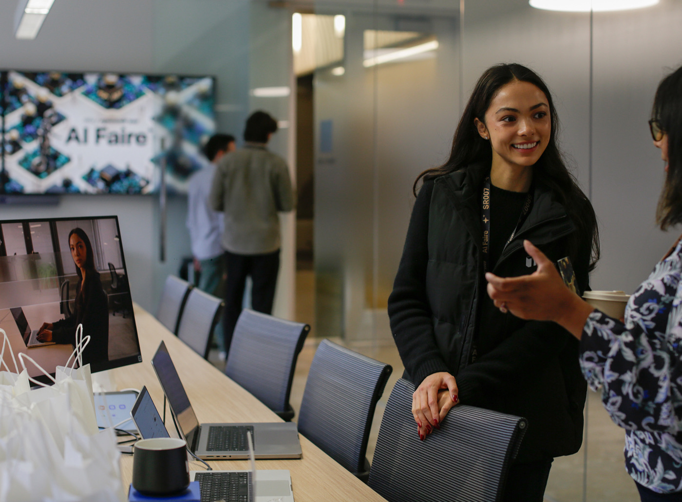 An AI Faire host smiling and talking with a guest next to a table of laptops